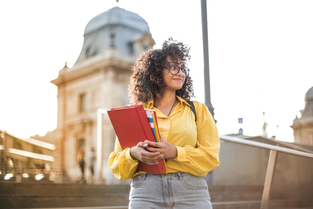 Studentin mit Heften in der Hand und gelben Bluse - Ratgeber Studieren in Spanien: Campus M University.