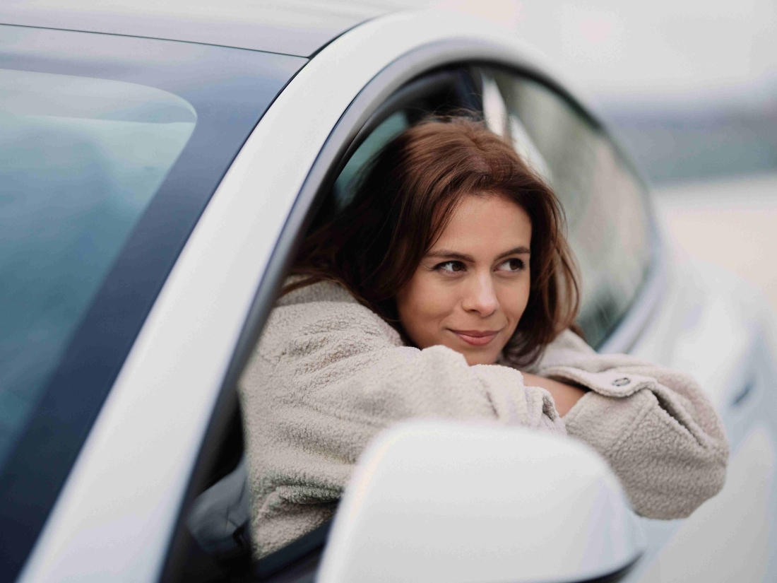 Frau im grauen Mantel lehnt aus einem weißen Auto – Informationen zum staatlich anerkannten Automobilwirtschaft Studium.