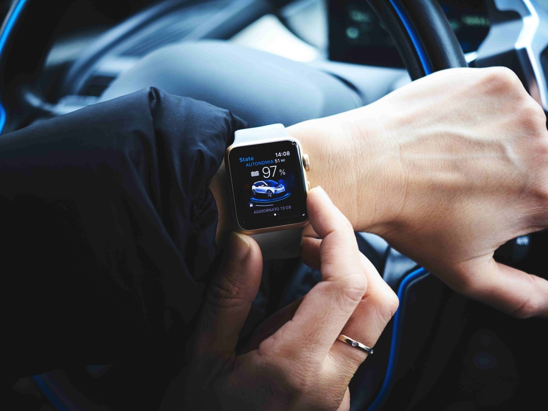 Eine Hand mit einer Apple Watch, auf der eine Auto-App angezeigt wird, während jemand im Auto sitzt.