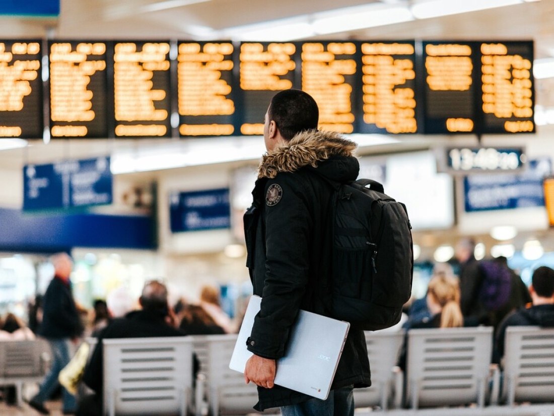 Junger Mann mit Laptop am Flughafen vor der Anzeigetafel - Ratgeber Studiumim Tourismus: Campus M University.