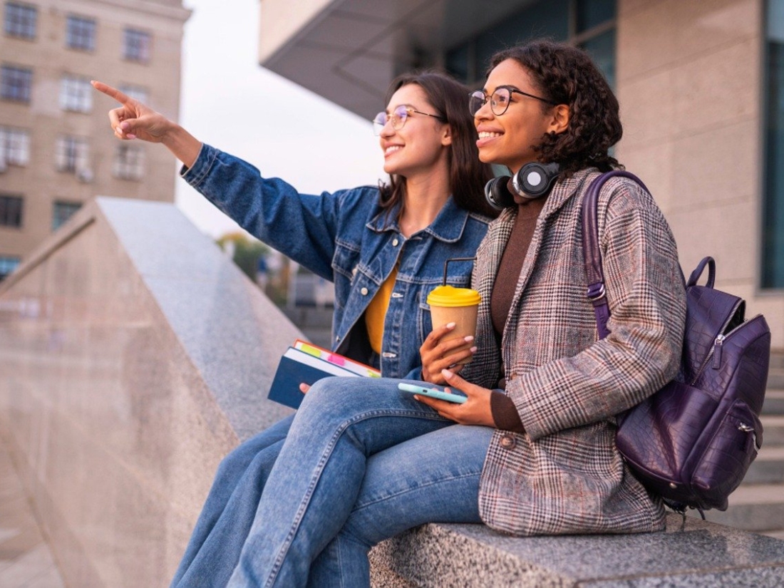 Zwei Studentinnen sitzen auf einem Treppengeländer, lachen miteinander und schauen in die Ferne. - Marketing Management Studium