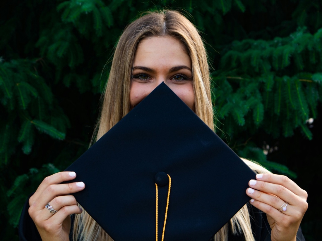 Glückliche junge Frau hält sich Abschlusskappe vor das Gesicht - Ratgeber Sportpsychologie Studium Campus M University