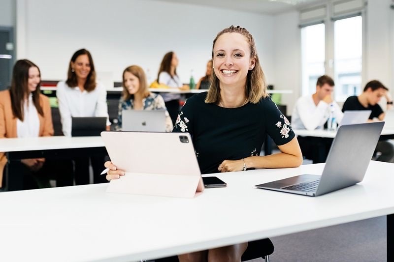 Glückliche Studentin am Tisch mit IPad - Ratgeber Campus M University Kosten.